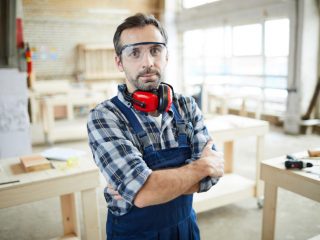 Carpenter in workshop wearing protective goggles and crossing arms on chest and looking at camera